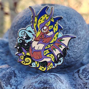 May include: A colorful enamel pin featuring a cartoon dragon with yellow, red, blue, and green accents. The dragon is surrounded by stars and clouds.