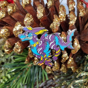 Könnte beinhalten: Eine violette und blaue Emaille-Anstecknadel mit einem Drachen mit gelben Akzenten. Der Drache steht auf einer Kiefernzapfen mit goldenem Glitzer.