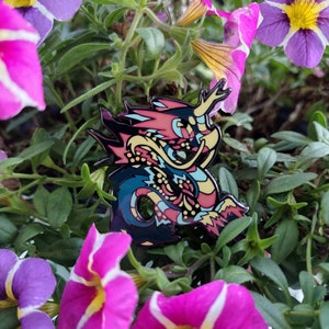 Puede incluir: Un pin de esmalte colorido con un dragón con escamas azules, rosas, amarillas y rojas. El dragón está enroscado con la cola en la boca. El pin está rodeado de flores rosas y moradas.