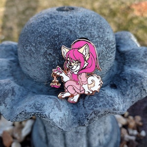 May include: A detailed enamel pin featuring a cartoon fox character with pink hair and clothing. The fox is holding a small, heart-shaped object. The pin has gold-colored metal accents and is set against a blurred outdoor background.