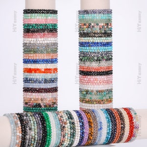 Könnte beinhalten: Eine Sammlung bunter Edelstein-Armbänder, die auf zwei weißen Ständern gestapelt sind. Die Armbänder bestehen aus kleinen, runden Perlen in verschiedenen Farben, darunter Blau, Grün, Rot, Pink und Schwarz.