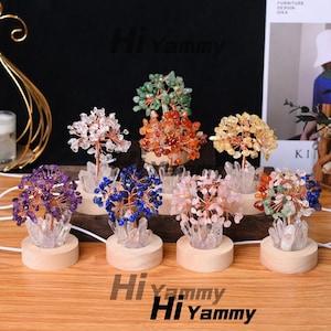 Op de afbeelding: Een verzameling decoratieve kristallen boomsculpturen in verschillende kleuren, waaronder paars, blauw, roze en groen. Elke boom heeft een draadgewikkeld ontwerp met kristallen accenten, gemonteerd op een houten basis. De bomen zijn verlicht.