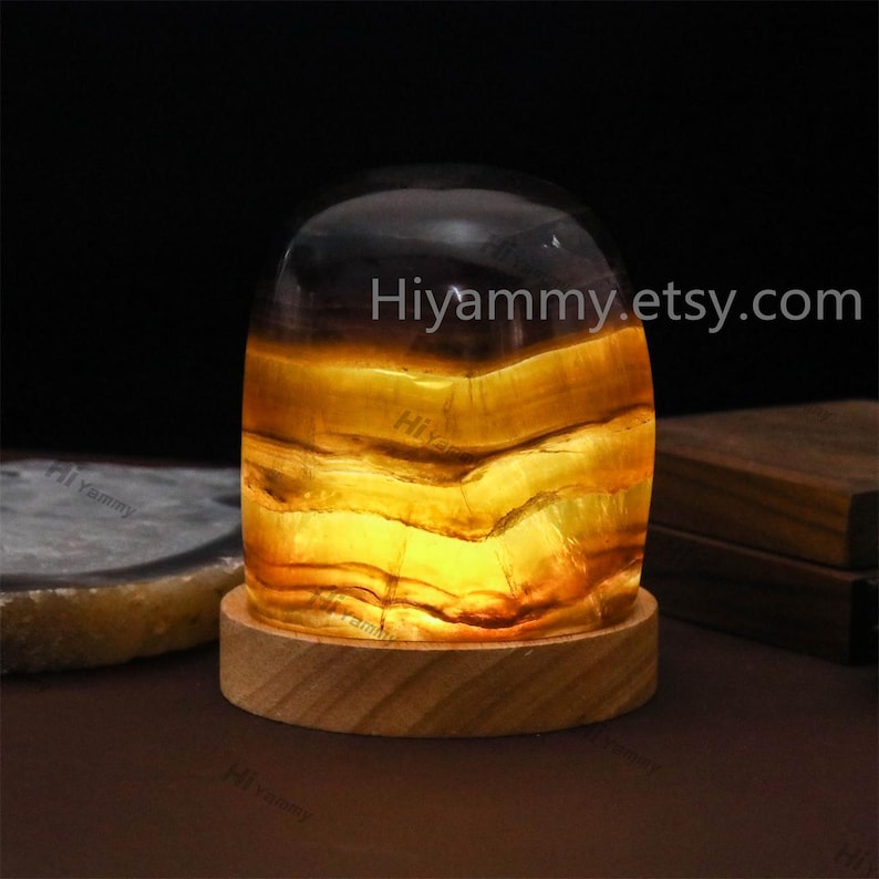 May include: A yellow and brown agate geode lamp with a wooden base. The geode is lit from within, creating a warm glow. The lamp is on a dark surface. Hiyammy.etsy.com