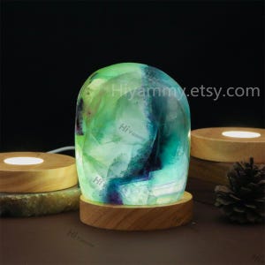 May include: A green and blue fluorite crystal lamp with a wooden base. The crystal is illuminated from within, creating a soft glow. The lamp is sitting on a wooden surface.
