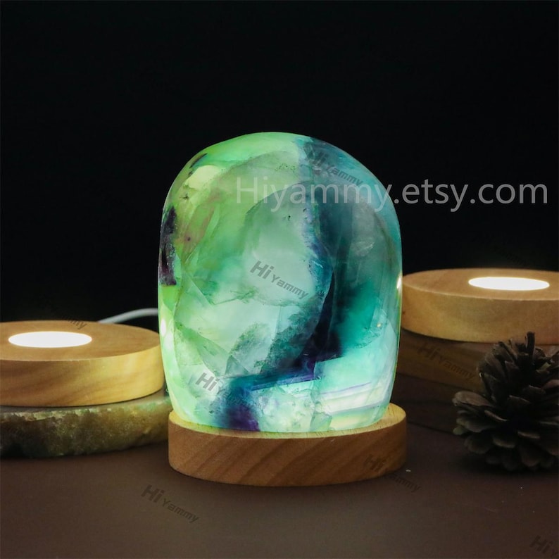 May include: A green and blue fluorite crystal lamp with a wooden base. The crystal is illuminated from within, creating a soft glow. The lamp is sitting on a wooden surface.