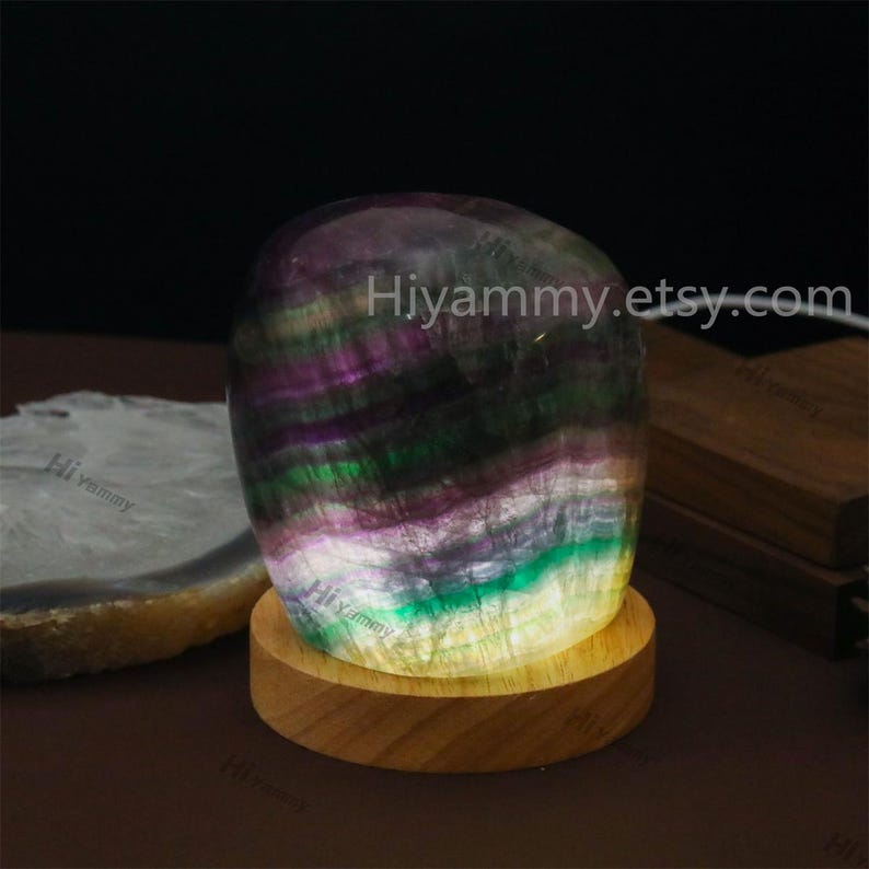 May include: A multi-colored fluorite crystal sphere with a wooden base and a warm white light emanating from the bottom. The crystal is a mix of purple, green, and yellow hues. The base is a natural wood color.