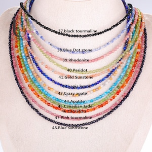 Peut inclure: Un collier composé de 11 rangs de perles de pierres précieuses de différentes couleurs. Les perles sont toutes petites et rondes, et sont enfilées sur un cordon fin. Le collier est présenté sur un fond blanc. Les rangs sont étiquetés avec le type de pierre précieuse, notamment la tourmaline noire, la pierre bleue à points, la rhodonite, le péridot, la pierre de soleil dorée, le lapis-lazuli, l'agate folle, l'apatite, la jadéite canadienne, la cornaline, la tourmaline rose et le grès bleu.