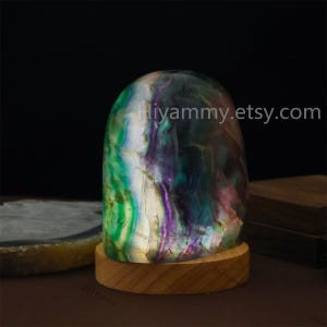 May include: A green, purple, and white fluorite crystal sphere on a wooden base. The crystal is illuminated from within, creating a glowing effect. The text "hiyummy.etsy.com" is visible in the image.
