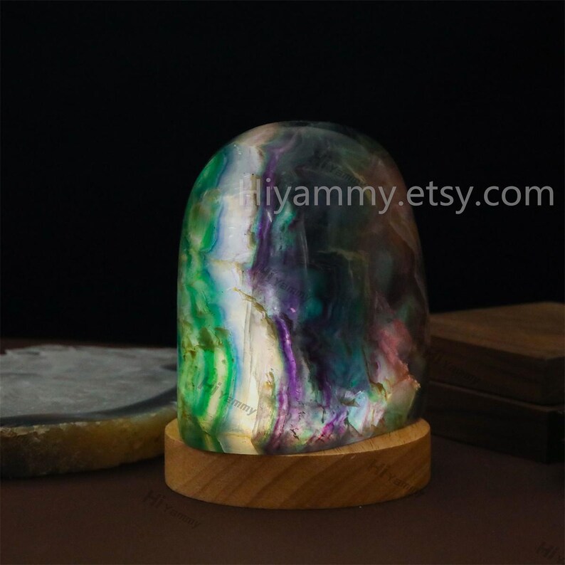 May include: A green, purple, and white fluorite crystal sphere on a wooden base. The crystal is illuminated from within, creating a glowing effect. The text "hiyummy.etsy.com" is visible in the image.