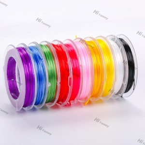 Könnte beinhalten: Ein Satz von zehn Spulen mit farbigem elastischem Garn, aufgewickelt auf transparente Kunststoffspulen. Die Farben sind lila, blau, grün, rot, rosa, gelb, orange, weiß und schwarz.