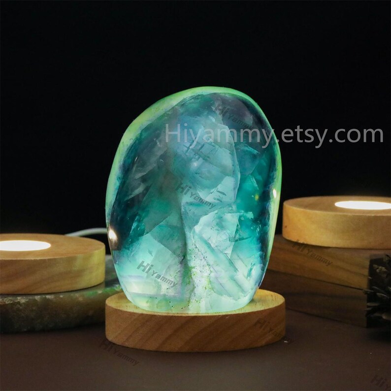 May include: A green and blue fluorite crystal ball on a wooden base with a light shining through it. The crystal ball is illuminated from the inside, creating a glowing effect. Hiyammyetsy.com is printed on the base.
