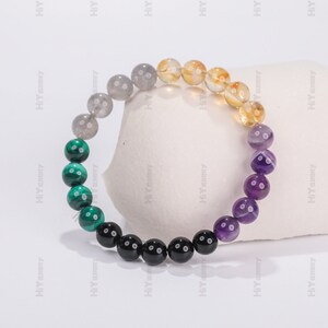 May include: A beaded bracelet with seven different colored stones. The bracelet is made of gray, green, black, purple, and yellow beads.