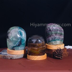 May include: Three polished fluorite crystal spheres on wooden bases. The crystals are green, purple, and brown with a layered, striated pattern. The bases are made of light brown wood.