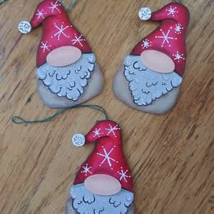 Pode incluir: Três ornamentos de gnomo de madeira feitos à mão com chapéus vermelhos e flocos de neve brancos. Cada gnomo tem uma barba branca e um rosto rosa.