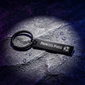 May include: A black metal rectangular keychain featuring a split ring and a flat tag. The tag is engraved with the text "PRINCESS POSSE" in a white serif font, accompanied by a small graphic of a cat wearing glasses and a crown.