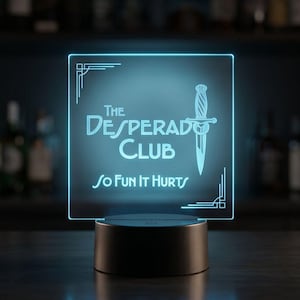 May include: A glowing blue LED sign with the text "The Desperado Club" and "So Fun It Hurts" displayed on a square panel. A dagger graphic is centered above the text. The sign is mounted on a black base, suitable for a bar or entertainment space.