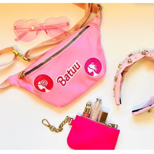 May include: Pink fanny pack with a zipper closure and gold hardware. The fanny pack has three pink embroidered patches, one with the word "Batuu" and two with a female silhouette. The fanny pack is laying on a white surface with a pair of pink heart-shaped sunglasses, a pink headband with pearl accents, and a small pink pouch with a gold chain.