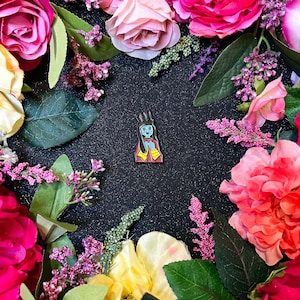 May include: A blue and red enamel pin of a cartoon character with a crown and a blue face. The pin is surrounded by pink and white flowers.