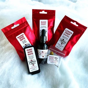 May include: An assortment of products from Wild Art Farm, including red pouches labeled "Electric Love Nuts," "Buzz Dust," and "Lemon Drop Spilanthes." A black bottle of "Spilanthes 2x Tincture" and a small container of "Freaks Electric Buzz Buds" are also visible.