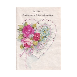 Könnte beinhalten: Vintage Valentinstag-Geburtstagskarte. Die Karte zeigt ein herzförmiges Design mit einem Blumenarrangement aus rosa und roten Rosen, blauen Blumen und grünen Blättern. Die Karte ist mit einer Spitzenborte und rosa Bändern verziert. Der Text "For Your Valentine's Day Birthday" steht oben.