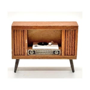 Puede incluir: Un mueble de tocadiscos en miniatura de madera de estilo moderno de mediados de siglo. El mueble marrón alberga un tocadiscos blanco. El mueble tiene patas negras y una rejilla de altavoz con dos detalles circulares. Aproximadamente 15 cm de ancho.