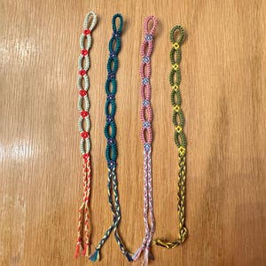 May include: Four colourful friendship bracelets. The bracelets are in red and white, teal and blue, pink and blue, and yellow and green. Each has a woven design with a loop at the top and braided ties at the bottom, displayed on a wooden surface.