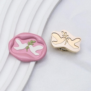 Könnte beinhalten: Ein rosa Wachssiegel und eine Holzbrosche mit einem Taubendesign. Das Wachssiegel hat eine rosa Basis mit zwei weißen Tauben, die einen grünen Olivenzweig halten. Die Holzbrosche hat eine helle Holzfarbe mit einer goldenen Umrandung der Tauben und des Olivenzweigs.