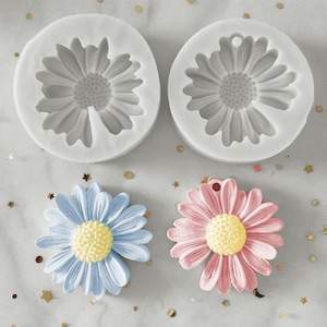 May include: Two silicone moulds for making daisy-shaped decorations. The moulds are white and have a daisy design. There are two finished daisies, one blue and one pink, next to the moulds.