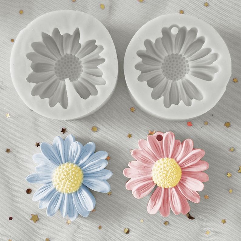 May include: Two silicone moulds for making daisy-shaped decorations. The moulds are white and have a daisy design. There are two finished daisies, one blue and one pink, next to the moulds.