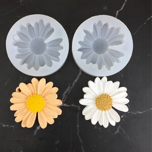 May include: Two silicone moulds for making daisy-shaped decorations. The moulds are white and have a daisy design. Two finished daisies are shown, one in white and one in peach.