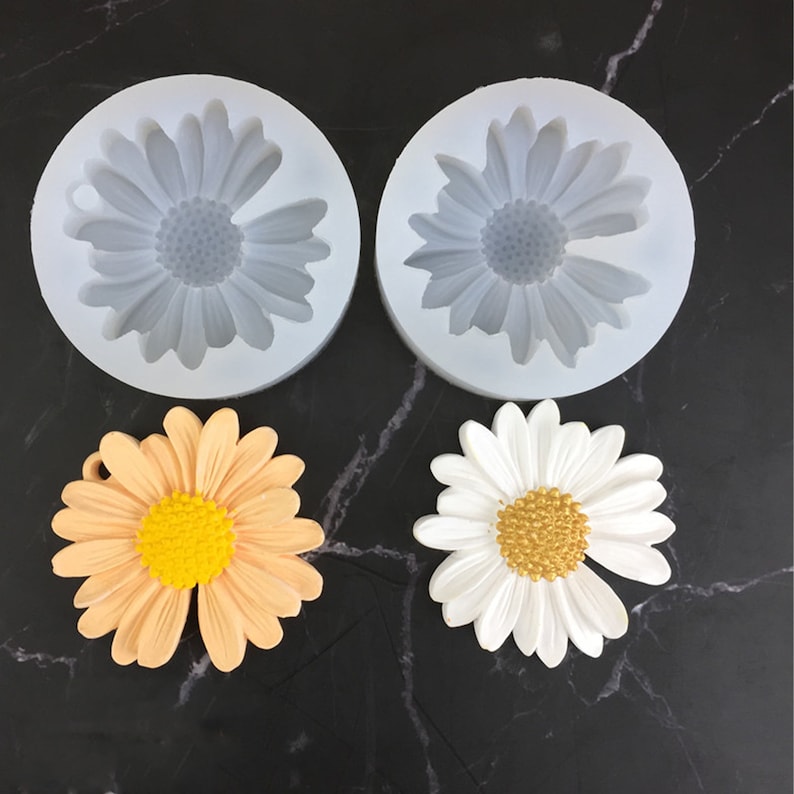 May include: Two silicone moulds for making daisy-shaped decorations. The moulds are white and have a daisy design. Two finished daisies are shown, one in white and one in peach.