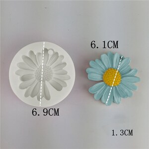 May include: A white silicone mould shaped like a daisy flower, measuring 6.9 cm in diameter. Next to it is a blue daisy flower made of resin, measuring 6.1 cm in diameter and 1.3 cm in height.