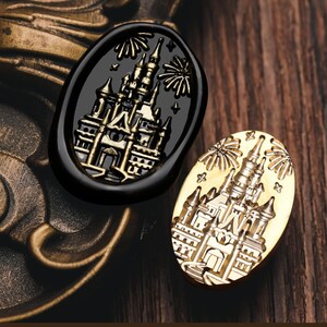 Pode incluir: Duas selos de cera ovais. Uma é preta com um desenho de castelo dourado e fogos de artifício. A outra é dourada com o mesmo desenho de castelo e fogos de artifício. Os selos estão em uma superfície de madeira.