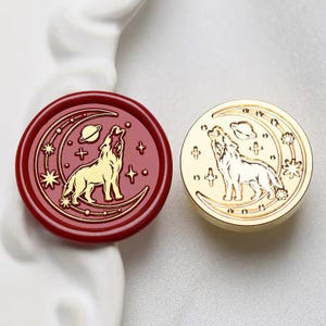 Könnte beinhalten: Zwei runde Wachssiegelstempel. Einer ist tiefrot mit einem goldenen Wolf, der den Mond, Sterne und einen Planeten anheult. Der andere ist goldfarben mit dem gleichen Design. Diese Stempel werden wahrscheinlich für dekorative Zwecke verwendet.