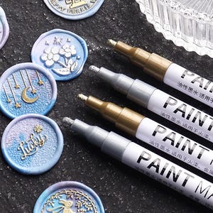 May include: Close-up of four paint markers with metallic gold and silver tips, alongside several wax seals with embossed designs. The markers have white barrels with black text. The wax seals are blue and purple with gold accents.