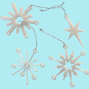 May include: Set of four white snowflake-shaped Christmas ornaments with silver string hangers. The ornaments are in a mid-century modern style.