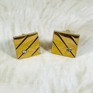 May include: A pair of gold cufflinks with a diagonal line pattern and a clear rhinestone in the centre of each.