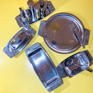 Könnte beinhalten: Ein Set silberner Servierplatten mit verzierten Rändern. Das Set enthält eine große runde Platte, zwei kleine rechteckige Platten und eine kleine ovale Platte. Die Platten sind auf einer gelben Oberfläche angeordnet.