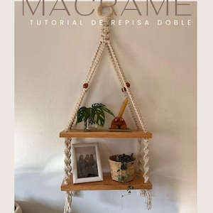May include: A macrame wall shelf with two wooden shelves. The shelf is made with natural coloured rope and has wooden beads. The shelves are decorated with a framed photo, a potted plant, and a small wooden figurine.