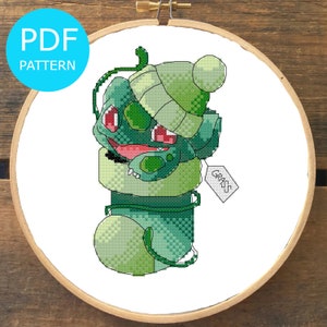 Puede incluir: Patrón de punto de cruz de un personaje Pokemon verde que lleva un gorro blanco y una bufanda verde. El personaje tiene ojos rojos y una etiqueta que dice "GRASS".