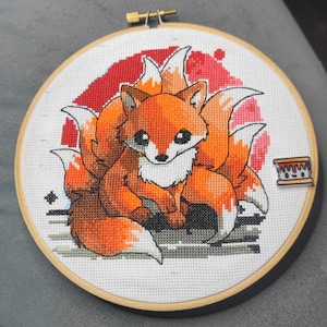 May include: Cross-stitch embroidery of a red fox with a nine-tailed kitsune design. The fox is sitting with its paws crossed and has a friendly expression. The background is a red and white watercolor-style design.