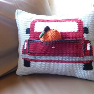 Puede incluir: Una almohada de crochet con forma de camioneta roja con una sola naranja en la caja. La almohada está en un sofá marrón.