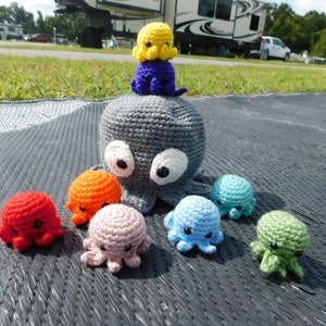 May include: A collection of crocheted octopus plushies in various colors, including gray, red, orange, pink, light blue, turquoise, and green. The largest octopus is gray and has white eyes. The smaller octopuses are arranged around the larger one.