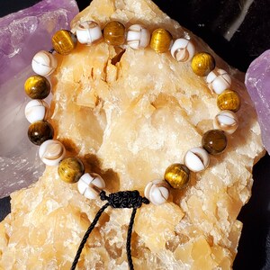 May include: A brown and white beaded bracelet with a black cord and tassel. The beads are made of tiger's eye and white shell.