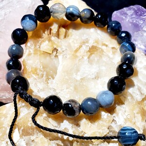 Op de afbeelding: Een armband van zwarte en grijze agaat met een zwarte koordsluiting. De armband is gemaakt van ronde kralen en wordt getoond op een lichtbruin oppervlak.