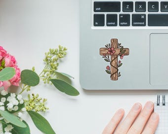 Puede incluir: Una cruz de madera con flores rosas y hojas verdes envueltas a su alrededor. La cruz está sobre un fondo gris claro.