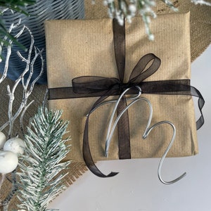 May include: A present wrapped in brown paper, tied with a black ribbon. A silver wire heart decoration sits on top. Winter greenery and a woven basket are in the background, creating a festive scene.