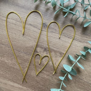 May include: Three gold wire heart shapes of varying sizes. The hearts are arranged on a wooden surface, with two larger hearts and one smaller heart. The wire is bent into the shape of a heart, with the ends of the wire crossed at the bottom.