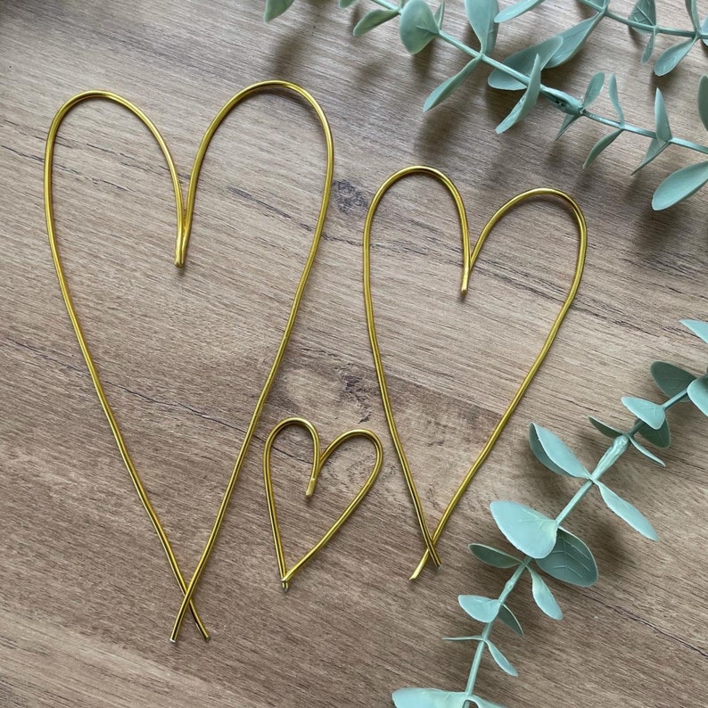 May include: Three gold wire heart shapes of varying sizes. The hearts are arranged on a wooden surface, with two larger hearts and one smaller heart. The wire is bent into the shape of a heart, with the ends of the wire crossed at the bottom.