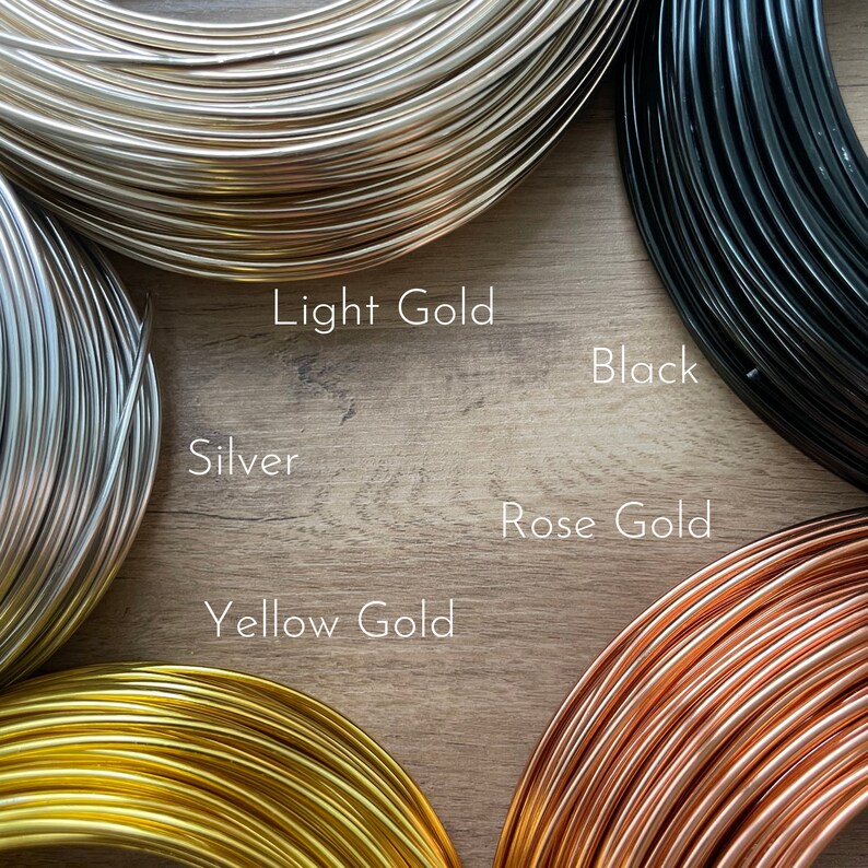 May include: Close-up of spools of wire in various colors, including light gold, silver, black, rose gold, and yellow gold. The image is taken on a wooden surface. Text labels identify each color.